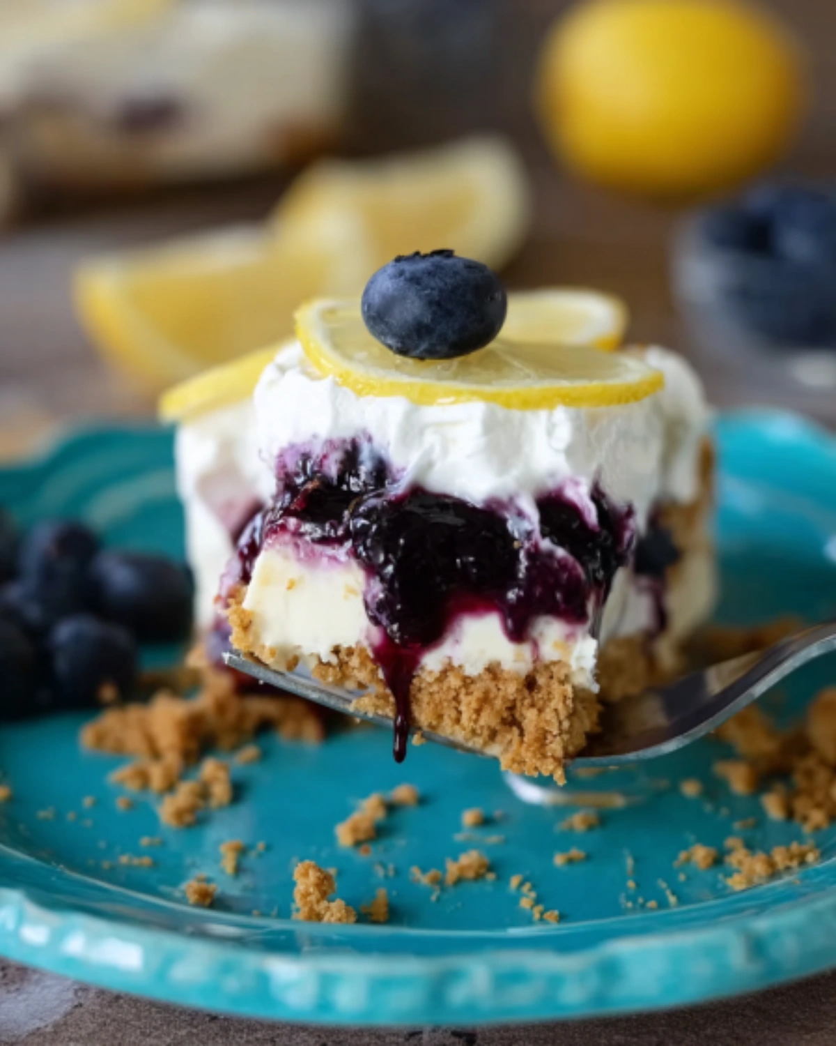 no-bake-lemon-blueberry-dessert-serving-on-plate Fork holding a slice of No Bake Lemon Blueberry Dessert with blueberry topping and lemon garnish on a turquoise plate