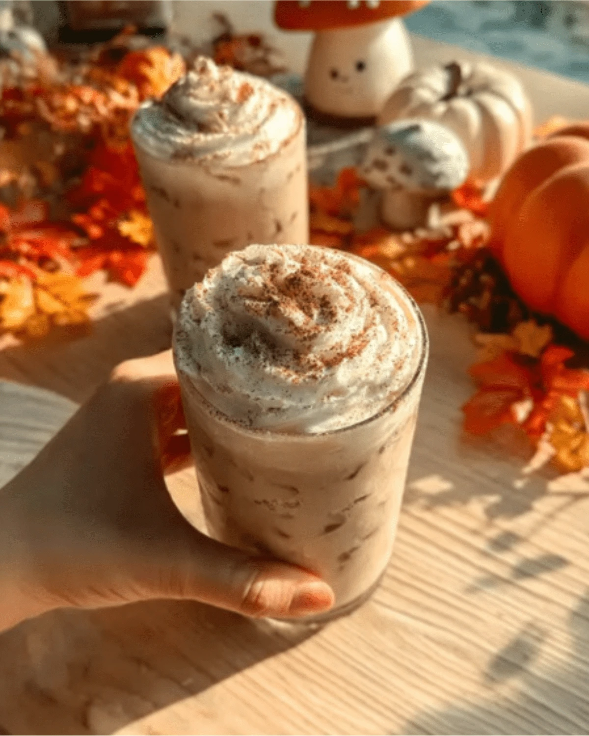 iced-pumpkin-cream-chai-autumn-vibes Hand holding a glass of iced pumpkin cream chai topped with whipped cream and cinnamon, surrounded by autumn pumpkins and leaves.