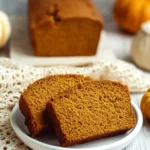Two slices of gluten free pumpkin bread on a white plate with the full loaf and pumpkins in the background