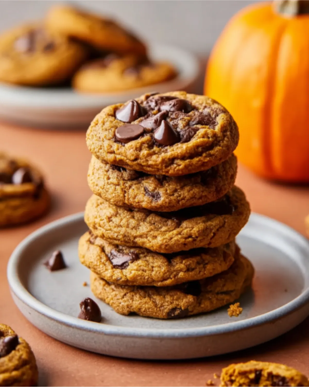 Stack of brown butter pumpkin chocolate chip cookies on a plate with chocolate chips and a pumpkin in the background.