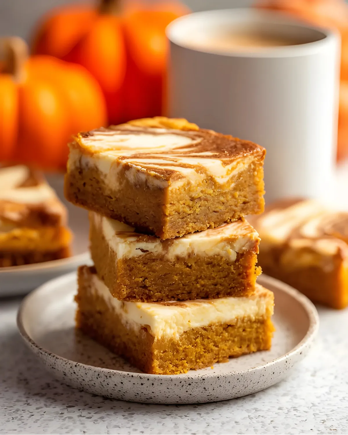 pumpkin-cheesecake-bread-swirled-bars Stacked pumpkin cheesecake bread bars with cream cheese swirls on a speckled plate beside coffee and pumpkins