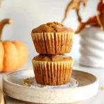 Two Kodiak Pumpkin Muffins stacked on a rustic ceramic plate with fall decor in the background.