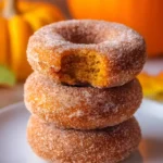 Pumpkin spice donuts stacked on a plate with a bite taken out of the top donut, surrounded by fall decorations.
