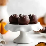 Pumpkin spice truffles topped with flaky sea salt displayed on a white cake stand, with mini pumpkins in the background.