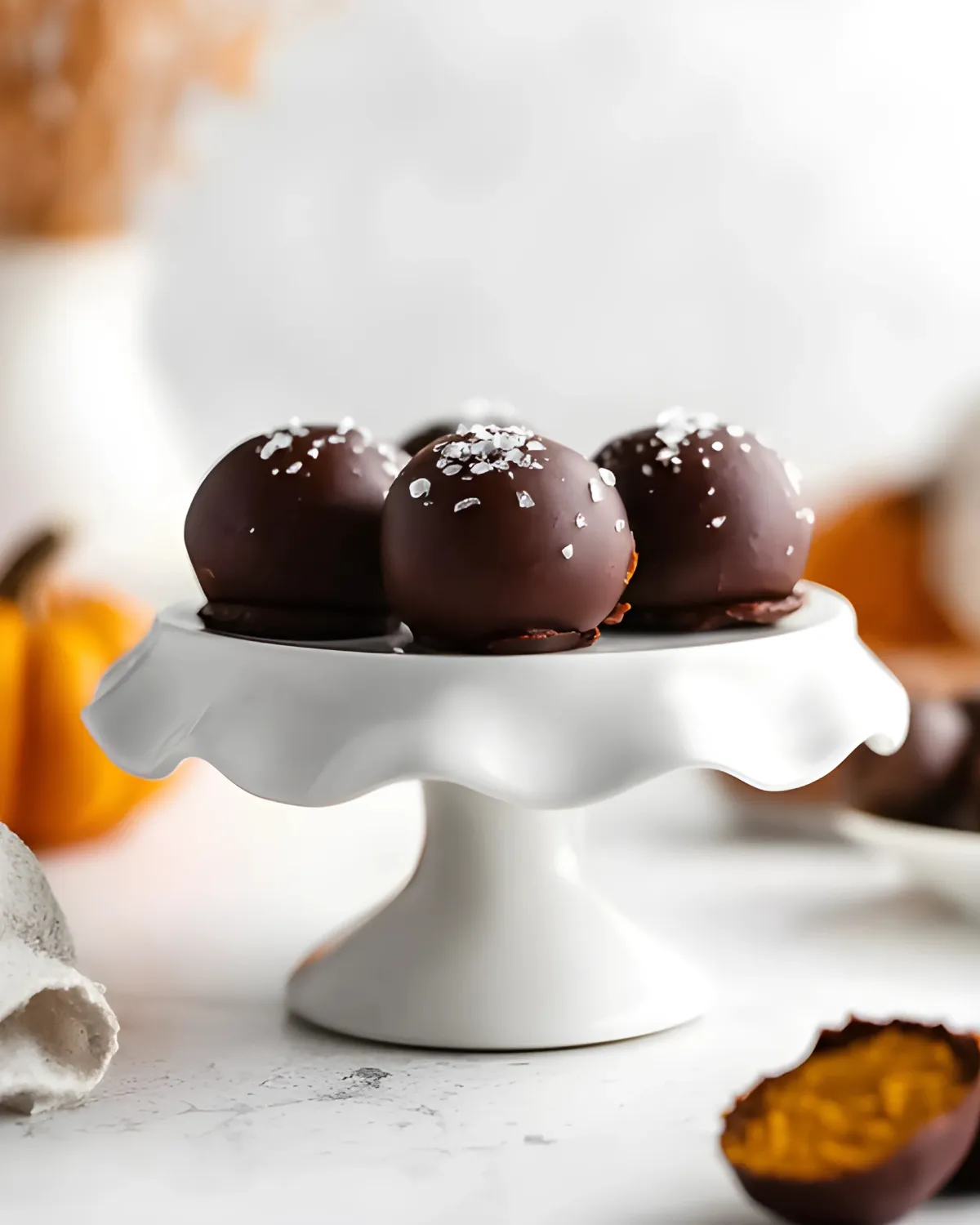 pumpkin-spice-truffles-on-white-cake-stand Pumpkin spice truffles topped with flaky sea salt displayed on a white cake stand, with mini pumpkins in the background.