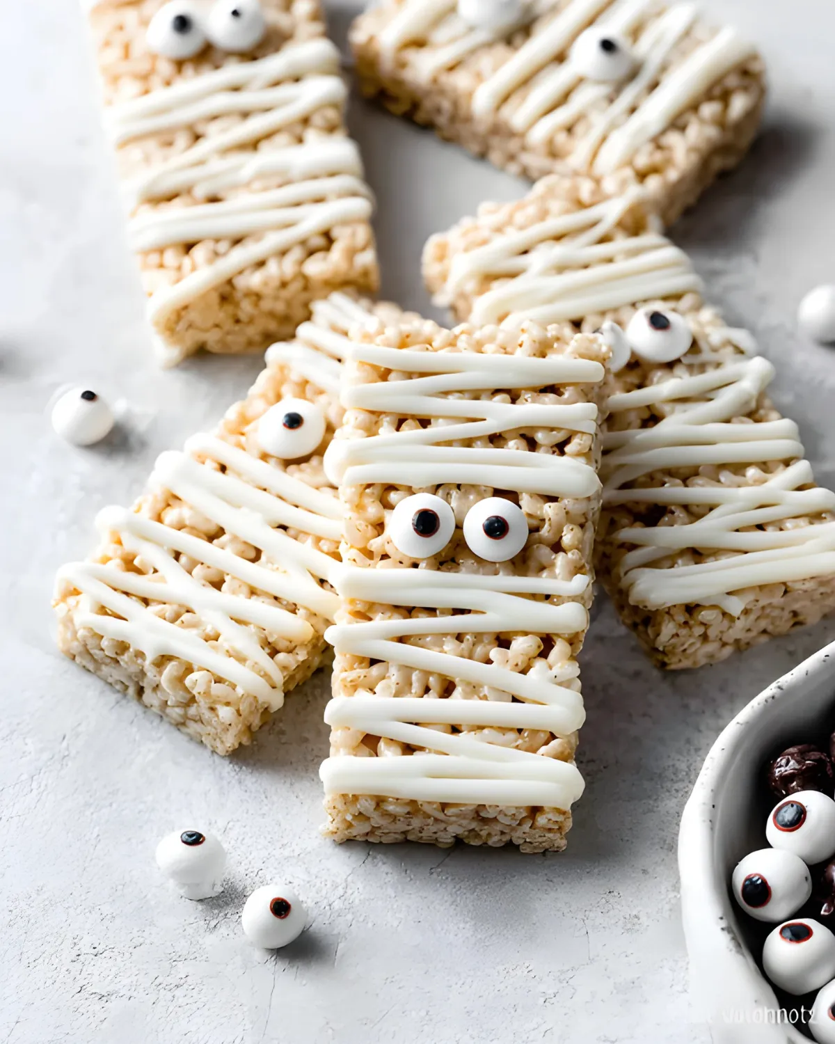 Halloween Rice Krispie Treats shaped like mummies with white frosting and candy eyes on a gray background
