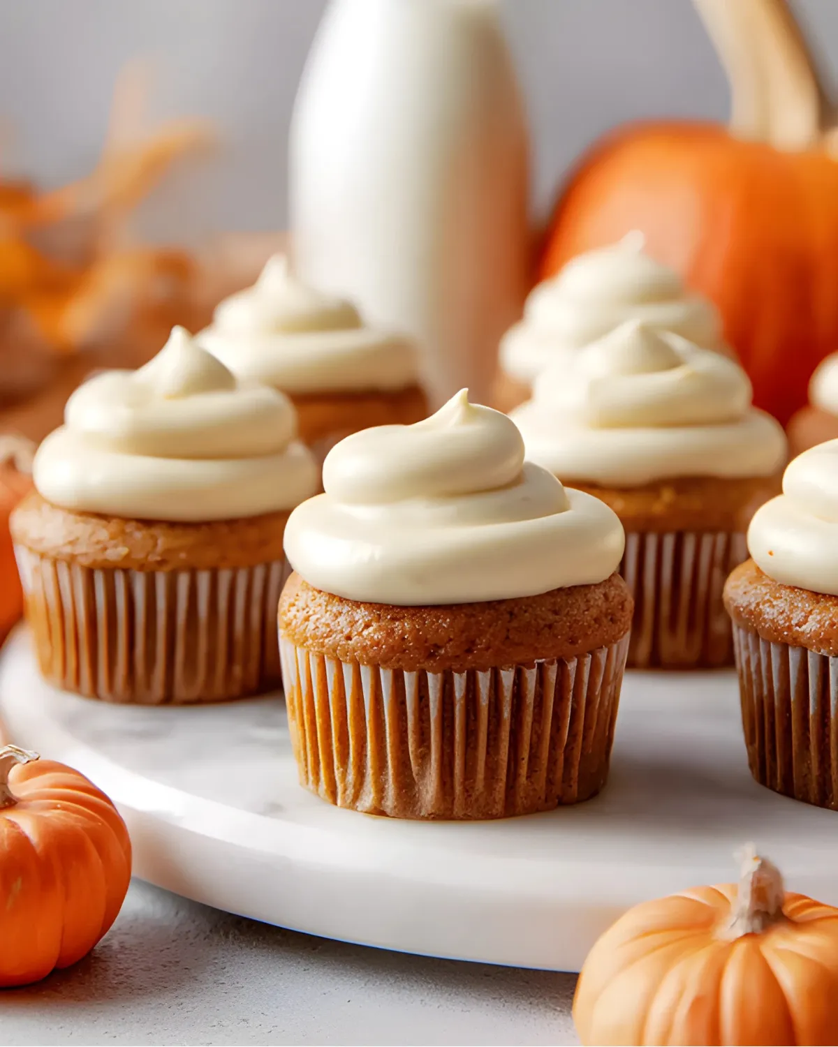 Pumpkin cupcakes topped with cream cheese frosting, surrounded by mini pumpkins and a bottle of milk