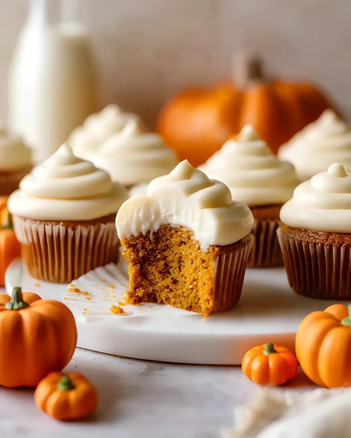 Pumpkin cupcakes with cream cheese frosting, one with a bite taken out, surrounded by mini pumpkins