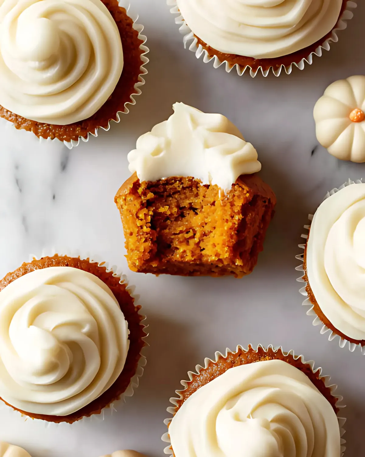 Top view of pumpkin cupcakes with cream cheese frosting, one cupcake with a bite taken out on a marble surface