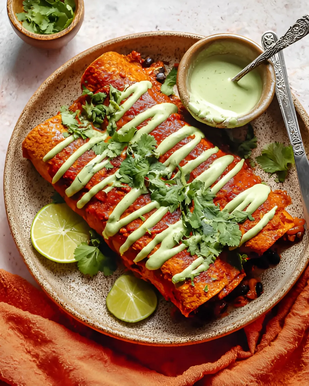 Pumpkin Black Bean Enchiladas topped with cilantro and creamy green sauce on a rustic plate with lime wedges
