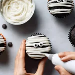 Hands decorating Halloween Mummy Cupcakes with white frosting and candy eyes