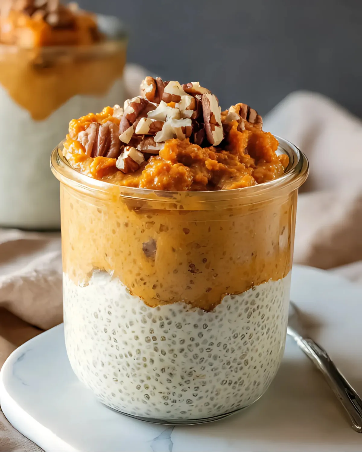 Pumpkin chia pudding layered in a glass jar, topped with chopped pecans on a marble plate.