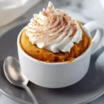 Pumpkin mug cake topped with whipped cream and cinnamon in a white mug on a gray plate