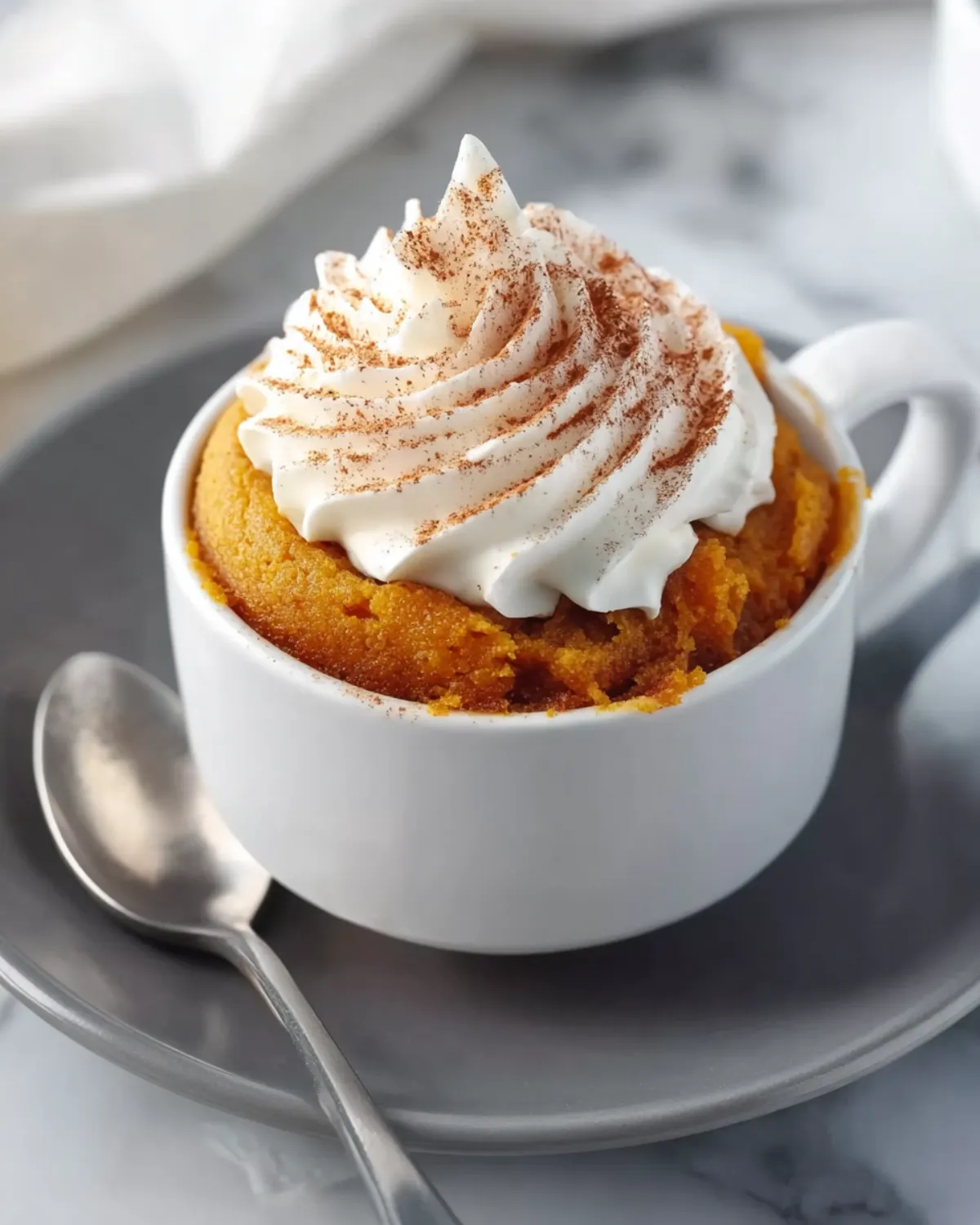 Pumpkin mug cake topped with whipped cream and cinnamon in a white mug on a gray plate