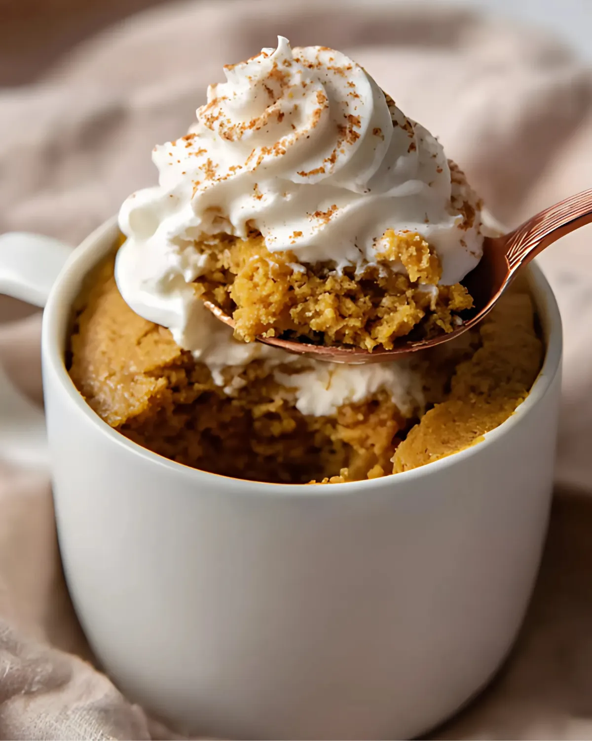 Pumpkin mug cake with a spoonful topped with whipped cream and cinnamon in a white mug