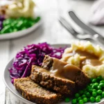 Sliced Vegan Mushroom Lentil Loaf topped with gravy, served with mashed potatoes, green peas, and red cabbage on a white plate.