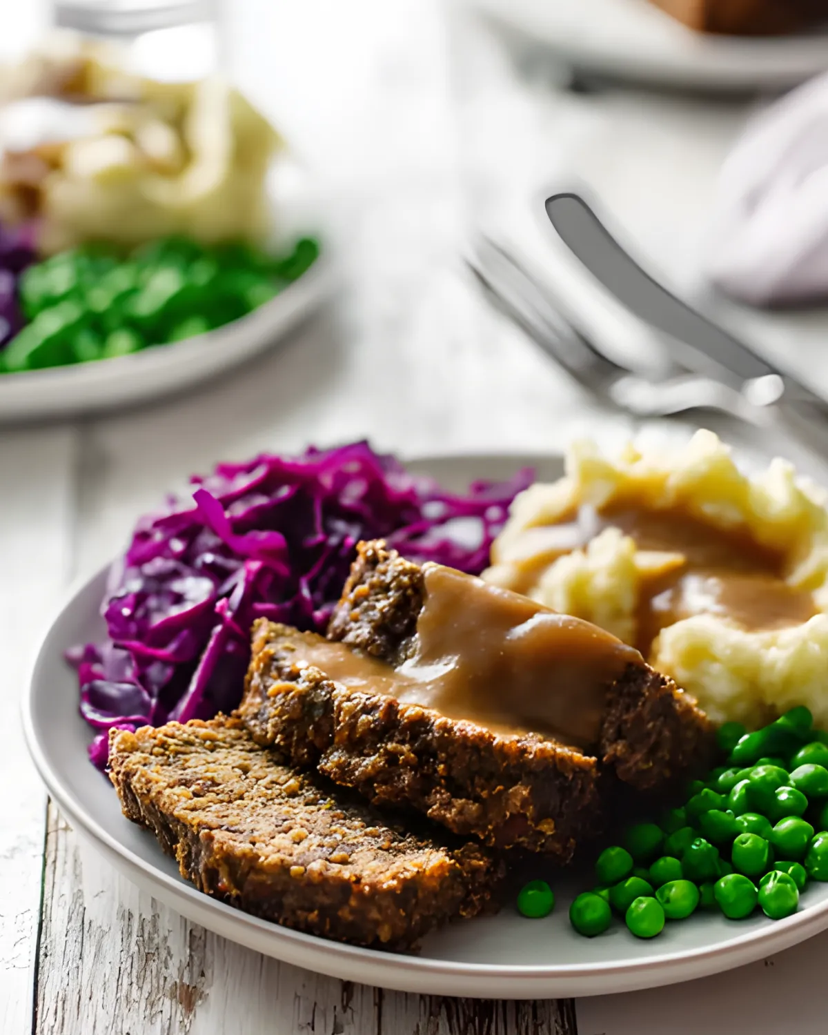Sliced Vegan Mushroom Lentil Loaf topped with gravy, served with mashed potatoes, green peas, and red cabbage on a white plate.