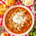 Bowl of crockpot chicken parmesan soup topped with shredded cheese and sour cream, served with garlic bread on a red checkered cloth.