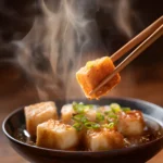 Agedashi Tofu with crispy golden exterior lifted by chopsticks over a bowl of savory dashi broth, garnished with sliced green onions and sesame seeds.