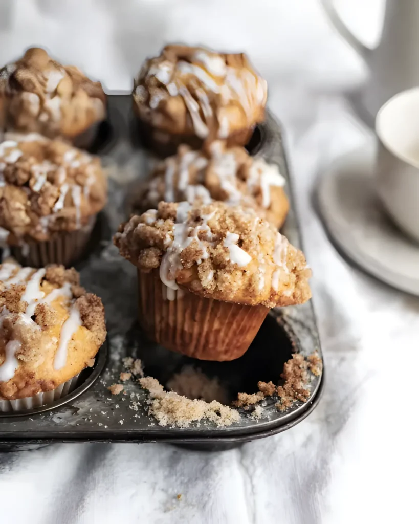 Tray of freshly baked coffee cake muffins with crumb topping and glaze, with one muffin lifted from the tin.