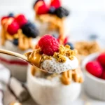 Spoonful of protein chia pudding with peanut butter, granola, and a fresh raspberry held above a glass jar dessert