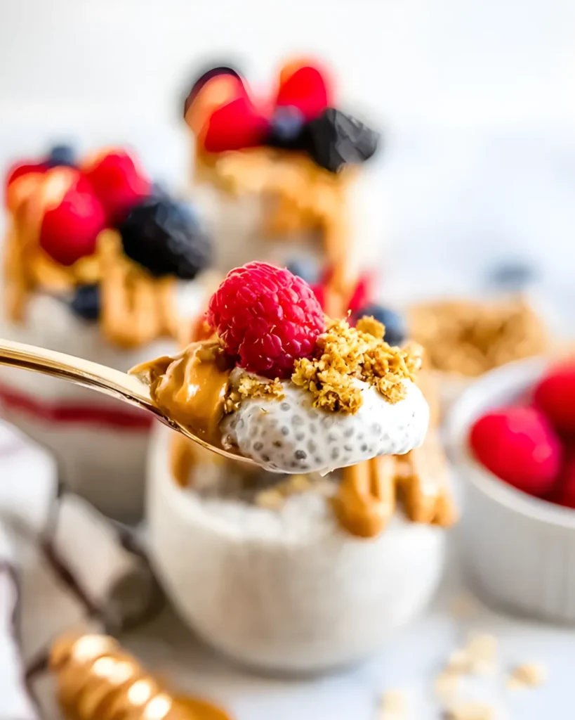 Spoonful of protein chia pudding with peanut butter, granola, and a fresh raspberry held above a glass jar dessert