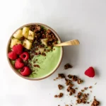 Tropical Matcha with Pineapple smoothie bowl topped with raspberries, pineapple chunks, chocolate granola, and a gold spoon