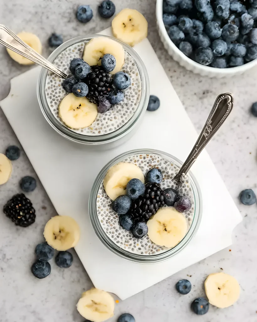 Two jars of vanilla chia pudding topped with banana slices, blueberries, and blackberries on a white board