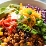 Taco Skillet served in a bowl with seasoned ground beef, black beans, shredded cheddar cheese, avocado slices, jalapeños, green onions, diced tomatoes, and purple cabbage.
